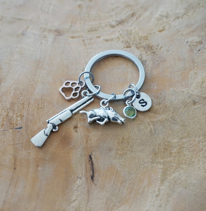 May include: Silver key chain with a variety of charms, including a paw print, a shotgun, a running boar, a green gemstone, and a silver charm with the letter 'S'.