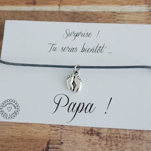 Peut inclure: Un bracelet en cordon gris avec un charme argenté de deux empreintes de pieds de bébé. Le bracelet est sur une carte blanche avec le texte "Surprise ! Tu seras bientôt... Papa !" et un petit cœur avec le texte "Handmade with Love".