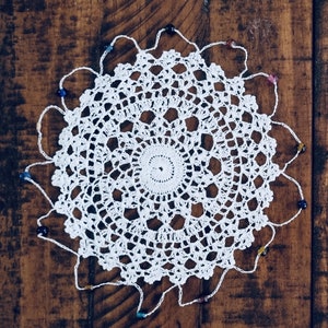 Pode incluir: Doily de crochê branco com um delicado padrão floral e uma borda de contas. O doily é circular e tem um furo central. As contas são de várias cores, incluindo azul, roxo e verde.