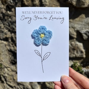 May include: A white card with the message "WE'LL NEVER FORGET YOU Sorry You're Leaving". It features a blue crocheted flower with a yellow centre, black stem and leaves. A leaving card.