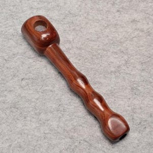 Pode incluir: Um cachimbo de madeira artesanal com acabamento castanho-avermelhado escuro. O cachimbo apresenta uma tigela cilíndrica com um pequeno orifício e um caule curvo e ergonómico. O caule tem uma série de reentrâncias arredondadas para a aderência.