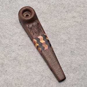 Puede incluir: Una pipa de madera marrón oscuro con un diseño geométrico en cobre y oro. Un diseño grabado es visible cerca del cuenco. La pipa está inclinada hacia abajo a la derecha sobre una superficie gris. La pipa mide unos 15 cm de largo.