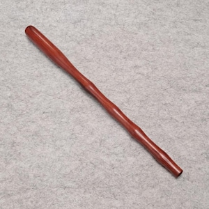 May include: A smooth, reddish-brown wooden rod with a tapered, cylindrical shape. The object features a polished finish and a consistent, slender profile that narrows slightly toward both ends, showcasing the natural grain of the wood material.