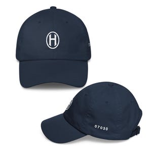 May include: A navy blue baseball cap featuring a white embroidered letter H inside an oval on the front panel. The side of the cap displays the white numerical text 07030, and it includes an adjustable strap closure at the back for a secure fit.