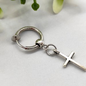 Op de afbeelding: Zilverkleurige clip-on oorbel met een kruisvormige hanger. De oorbel heeft een cirkelvormig ontwerp met een scharnierende sluiting. Het kruis heeft een eenvoudige, klassieke vorm.