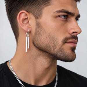 Pode incluir: Um brinco de tom prateado com dois pingentes retangulares finos de comprimentos diferentes suspensos por um pequeno aro circular. O brinco é usado no lóbulo da orelha e uma corrente de tom prateado é visível ao redor do pescoço.