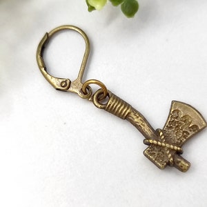 May include: An antique brass-toned metal earring featuring a small axe charm. The charm is attached to a lever-back closure by a jump ring. The axe design includes a textured blade and a wrapped handle detail, finished in a muted metallic tone.