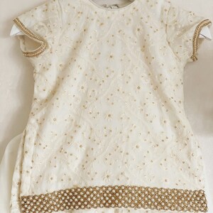 May include: A white embroidered baby girl's outfit with a pearl trim. The outfit includes a short-sleeved top and a skirt.