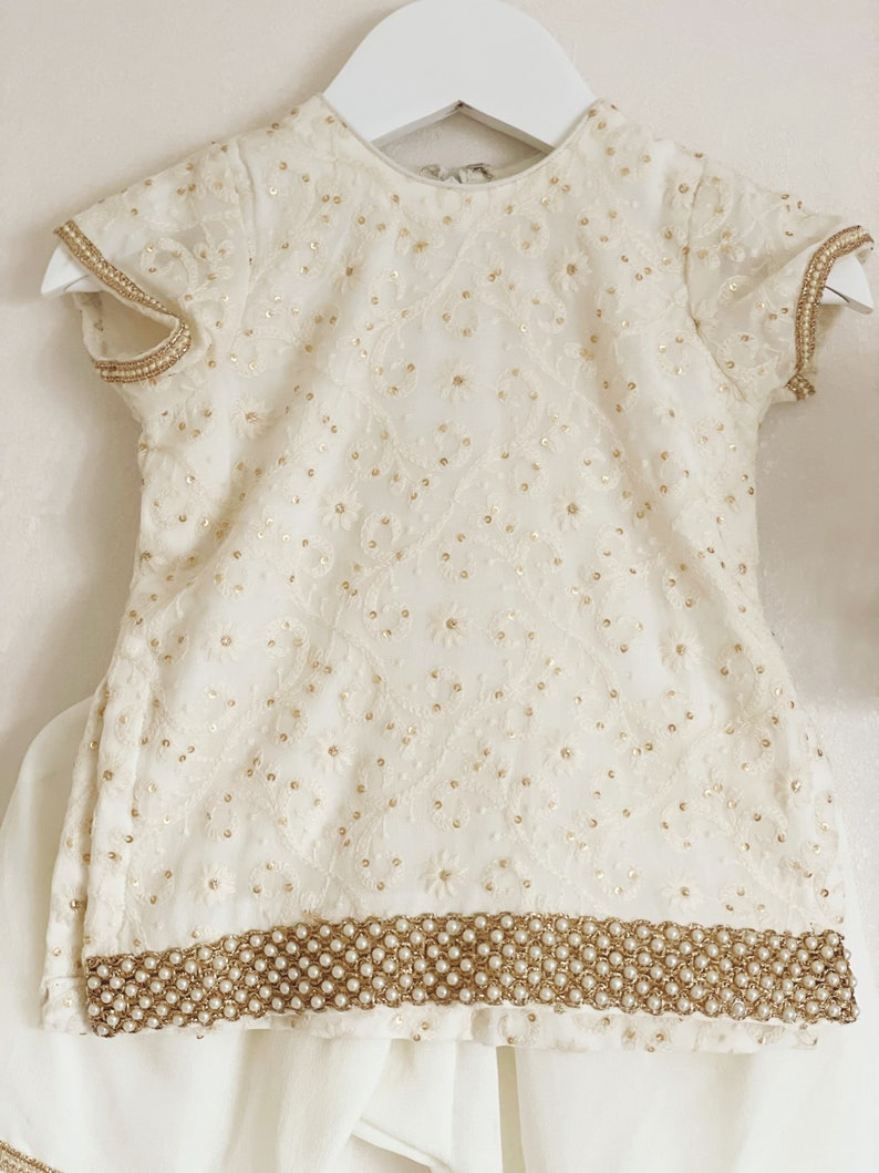 May include: A white embroidered baby girl's outfit with a pearl trim. The outfit includes a short-sleeved top and a skirt.