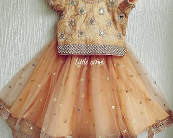 May include: A peach-colored girls' outfit with a gold embroidered top and a tulle skirt with gold sequins. The top has a round neckline and short sleeves. The skirt has a gold trim around the bottom. The outfit is labeled "Little orhni".