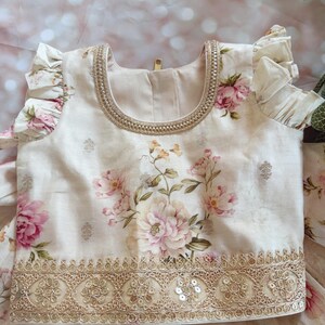 May include: Cream-colored floral top with pink and peach flowers. The top features a gold embroidered neckline and hem, with delicate ruffle sleeves. The fabric has a subtle sheen, and the design is detailed and elegant.