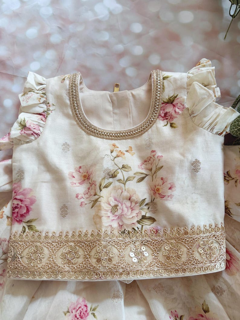 May include: Cream-colored floral top with pink and peach flowers. The top features a gold embroidered neckline and hem, with delicate ruffle sleeves. The fabric has a subtle sheen, and the design is detailed and elegant.
