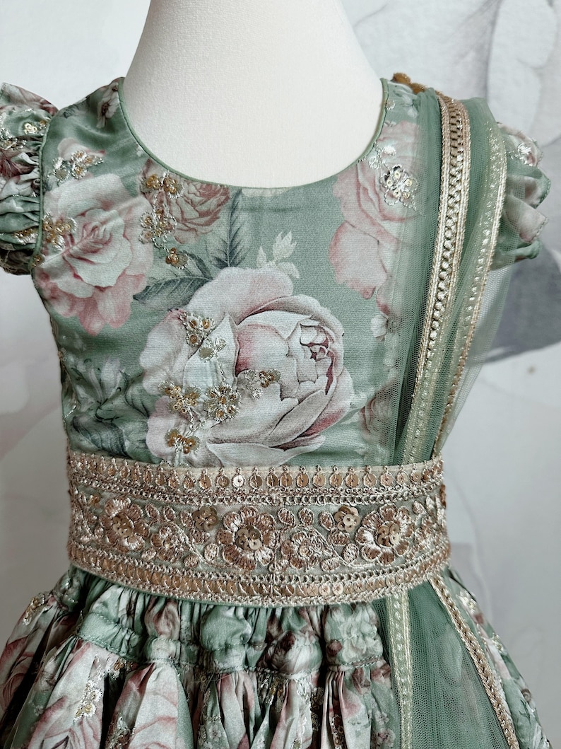 May include: A green and pink floral print dress with a gold embroidered belt and a matching sheer dupatta. The dress has a round neckline and short sleeves.