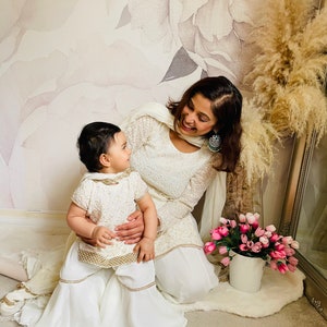 May include: A mother and daughter wearing matching white outfits with gold embroidery. The daughter is sitting on her mother's lap and they are both smiling at each other. The background is a floral wallpaper with a vase of pink flowers.