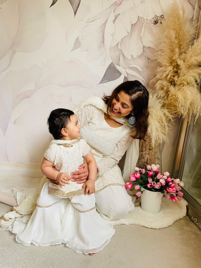 May include: A mother and daughter wearing matching white outfits with gold embroidery. The daughter is sitting on her mother's lap and they are both smiling at each other. The background is a floral wallpaper with a vase of pink flowers.