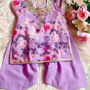 May include: A two-piece outfit for a child, in a lilac colour, featuring a floral print top with gold trim and matching trousers. The top has a decorative neckline with gold accents and mirrors.