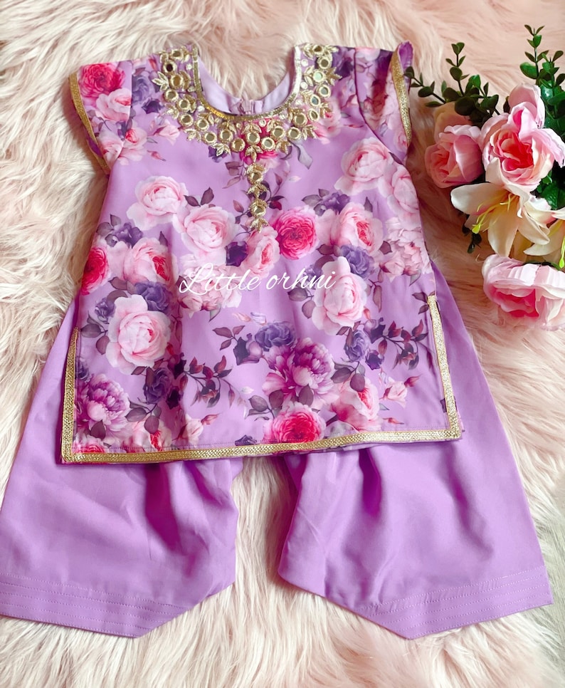 May include: A two-piece outfit for a child, in a lilac colour, featuring a floral print top with gold trim and matching trousers. The top has a decorative neckline with gold accents and mirrors.