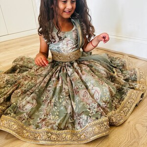 May include: A young girl wearing a sage green floral print dress with a gold embroidered trim and a gold belt. The dress has a gathered skirt and a fitted bodice. The girl is sitting on a wooden floor.