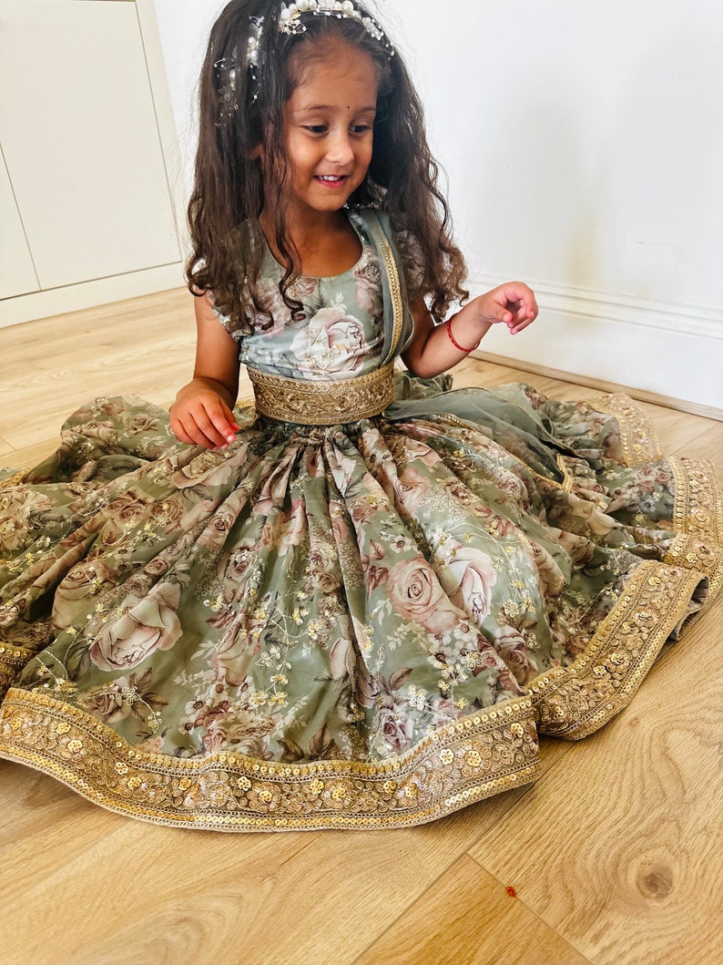 May include: A young girl wearing a sage green floral print dress with a gold embroidered trim and a gold belt. The dress has a gathered skirt and a fitted bodice. The girl is sitting on a wooden floor.