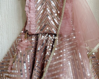 May include: A pink and gold sequined outfit for a young girl. The outfit includes a top with ruffled sleeves, a skirt, and a sheer dupatta. The outfit is decorated with sequins and mirror work.