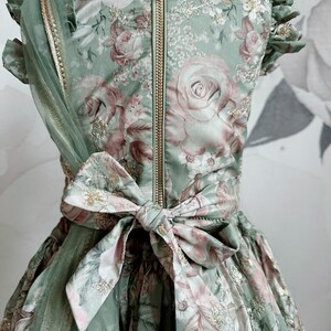 May include: A green floral dress with a gold zipper and a matching sash tied in a bow at the waist. The dress has a tulle overlay and gold embellishments.