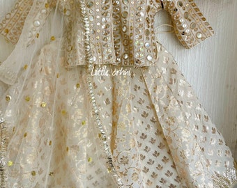 May include: A cream-colored girls' outfit with a matching skirt and top. The top is decorated with mirror work and the skirt has a floral pattern. The outfit is paired with a sheer dupatta with a gold border.