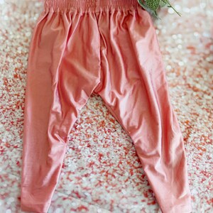 May include: A pair of peach-colored leggings with an elastic waistband. The leggings are made of a soft, flowing fabric and have a relaxed fit. The pants are displayed on a glittery surface.