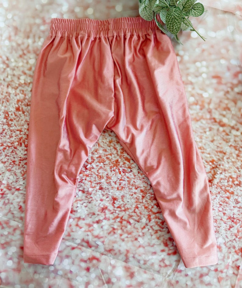 May include: A pair of peach-colored leggings with an elastic waistband. The leggings are made of a soft, flowing fabric and have a relaxed fit. The pants are displayed on a glittery surface.