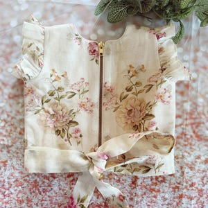 May include: Cream-colored top with a floral pattern in shades of pink, peach, and green. The top features a gold zipper, ruffled sleeves, and a tie at the waist. The floral design includes large peony-like blooms.
