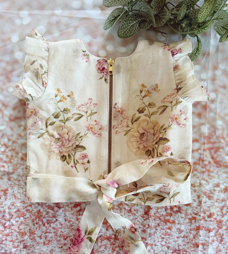 May include: Cream-colored top with a floral pattern in shades of pink, peach, and green. The top features a gold zipper, ruffled sleeves, and a tie at the waist. The floral design includes large peony-like blooms.