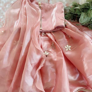 May include: A light pink dress with a flowing skirt and a bodice with a gold zipper. The dress is adorned with floral embellishments and a pearl trim. The fabric has a shiny, satin-like appearance, suggesting a formal occasion.