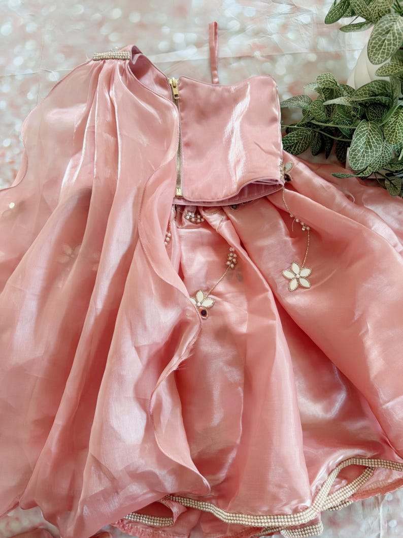 May include: A light pink dress with a flowing skirt and a bodice with a gold zipper. The dress is adorned with floral embellishments and a pearl trim. The fabric has a shiny, satin-like appearance, suggesting a formal occasion.