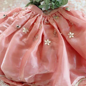 May include: A light pink skirt with a shimmering, translucent fabric. The skirt is adorned with white floral appliqu&eacute;s, small mirrors, and delicate pearl accents. The hemline is trimmed with a band of pearls. The skirt appears to be a child's garment.