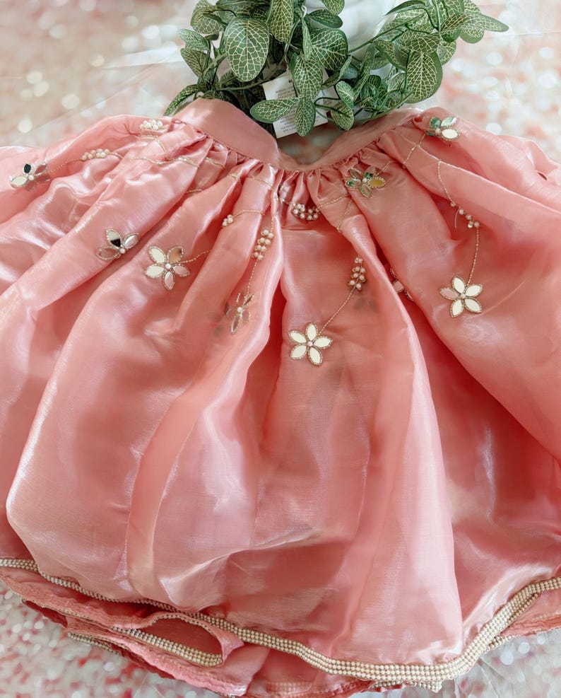 May include: A light pink skirt with a shimmering, translucent fabric. The skirt is adorned with white floral appliqu&eacute;s, small mirrors, and delicate pearl accents. The hemline is trimmed with a band of pearls. The skirt appears to be a child's garment.