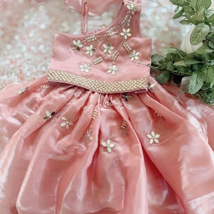 May include: A pink, one-shoulder dress with a layered skirt and floral embellishments. The bodice is adorned with white and silver flowers and a beaded trim. A matching pink scarf is draped over the dress, creating a formal look.