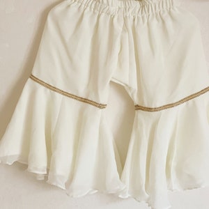 May include: White flared pants with a gold trim around the bottom. The pants have a wide leg and are made of a lightweight fabric.