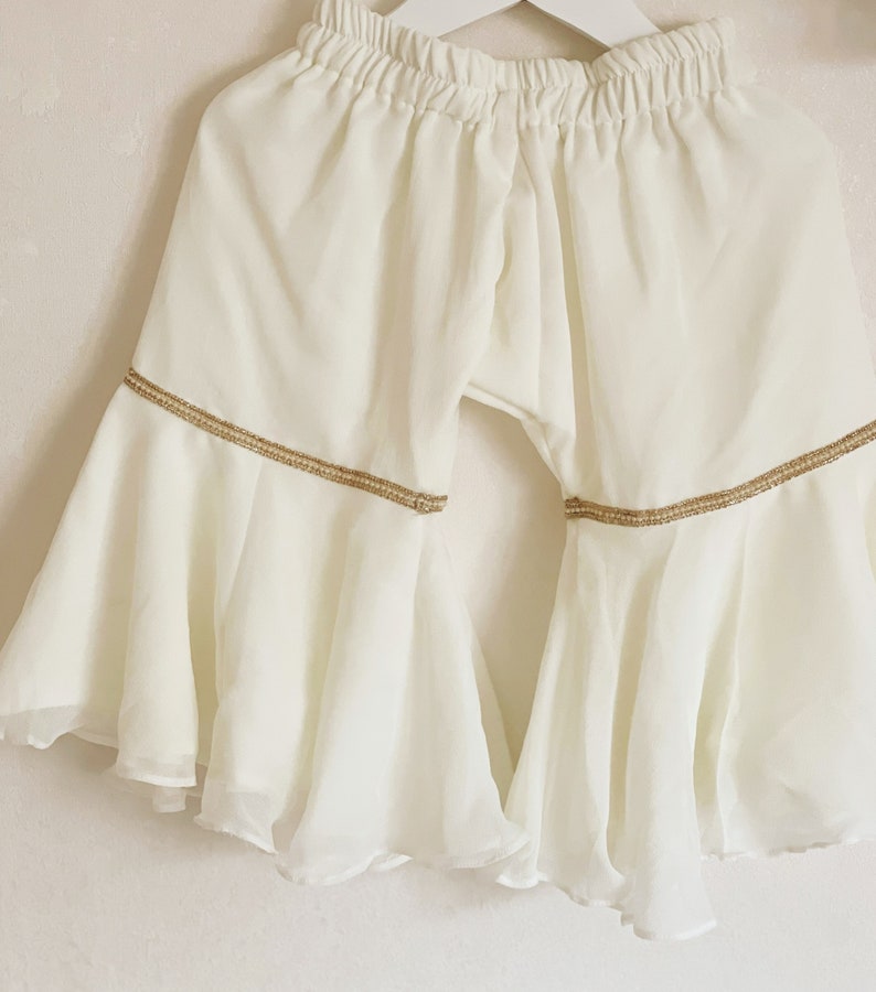 May include: White flared pants with a gold trim around the bottom. The pants have a wide leg and are made of a lightweight fabric.