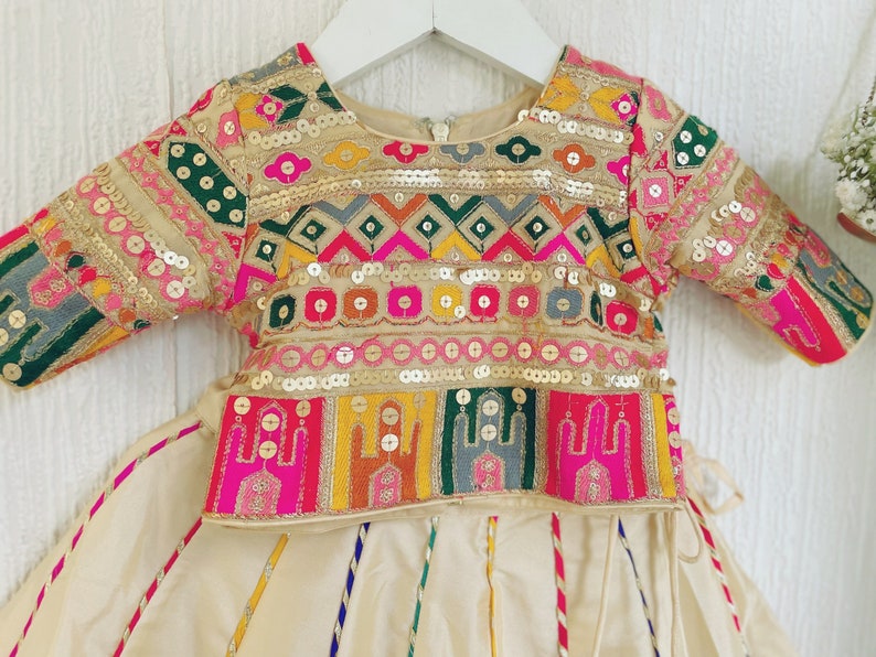 May include: A cream-colored, embroidered outfit for a child, featuring a top and skirt. The top has a round neckline and is adorned with colorful geometric patterns, sequins, and embroidery. The skirt is cream-colored with vertical stripes.