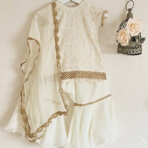 May include: A white outfit for a young girl, featuring a short-sleeved top with gold embroidery and a matching skirt with a ruffled hem. The outfit includes a sheer white dupatta with gold trim.