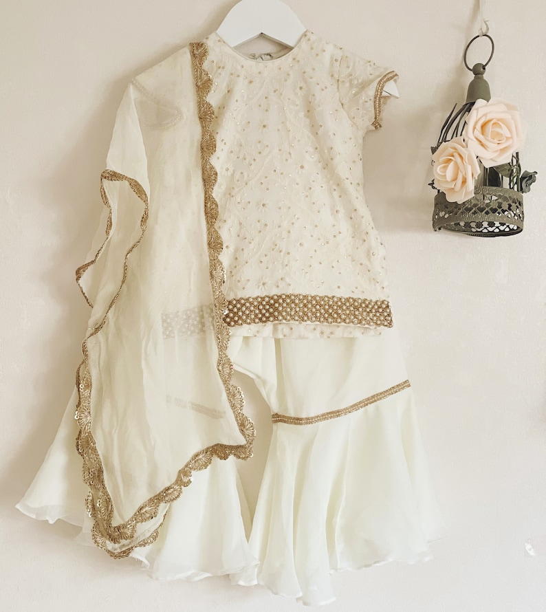 May include: A white outfit for a young girl, featuring a short-sleeved top with gold embroidery and a matching skirt with a ruffled hem. The outfit includes a sheer white dupatta with gold trim.