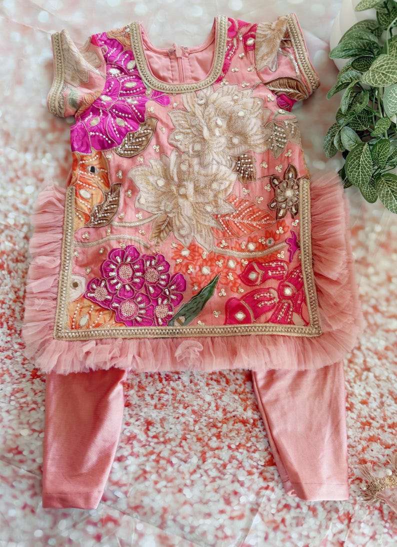 May include: Pink embroidered baby outfit with floral and sequin details. The top has short sleeves and a ruffled hem. The matching pants are a solid pink. The outfit is decorated with gold trim.