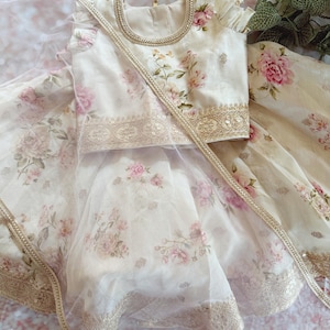 May include: A cream-colored floral print outfit featuring a top and skirt. The top has a round neckline and short sleeves. The skirt is layered with a sheer overlay. The outfit is adorned with gold trim and a matching sash. The floral pattern includes pink and peach roses.