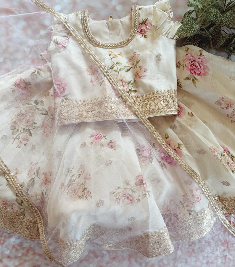 May include: A cream-colored floral print outfit featuring a top and skirt. The top has a round neckline and short sleeves. The skirt is layered with a sheer overlay. The outfit is adorned with gold trim and a matching sash. The floral pattern includes pink and peach roses.