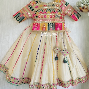 May include: A cream-colored, embroidered skirt and top set. The top is adorned with gold sequins and colorful embroidery. The skirt features vertical stripes in various colors and a decorative hem. The set includes matching decorative tassels.
