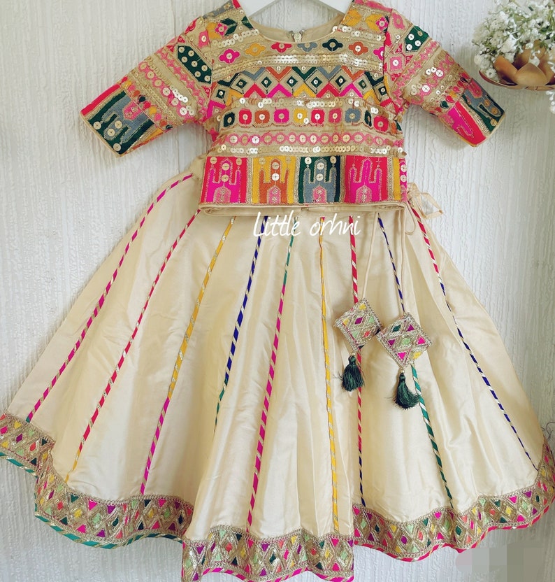 May include: A cream-colored, embroidered skirt and top set. The top is adorned with gold sequins and colorful embroidery. The skirt features vertical stripes in various colors and a decorative hem. The set includes matching decorative tassels.