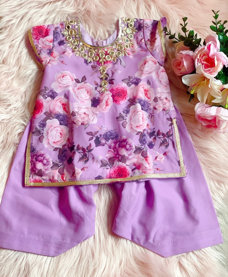May include: A purple floral print baby girl's outfit with a gold trim and a matching pair of trousers. The top has a decorative neckline with gold accents and mirror embellishments.