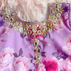 May include: A purple floral fabric with a gold and silver embellishment around the neckline. The embellishment features small round mirrors and leaf-shaped designs.