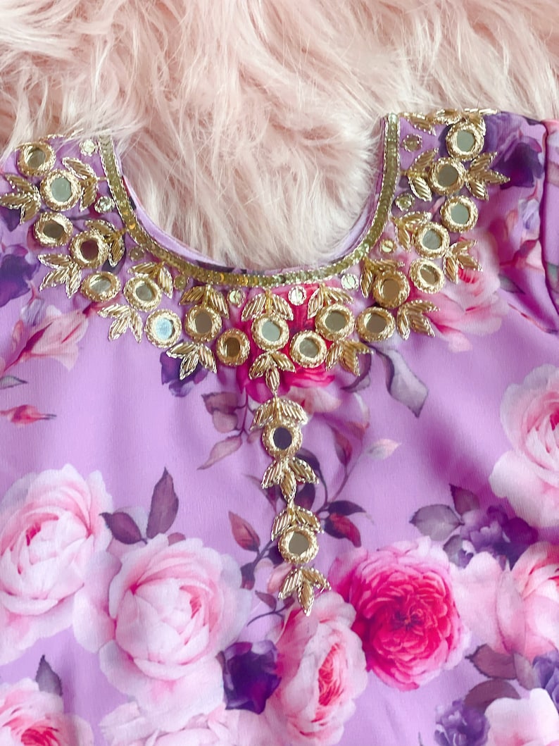 May include: A purple floral fabric with a gold and silver embellishment around the neckline. The embellishment features small round mirrors and leaf-shaped designs.