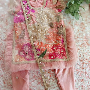 May include: A pink embroidered outfit for a child, featuring a top and pants. The top has intricate floral embroidery in shades of pink, orange, and gold, with a ruffled trim. A decorative trim with tassels and pearls accents the outfit.