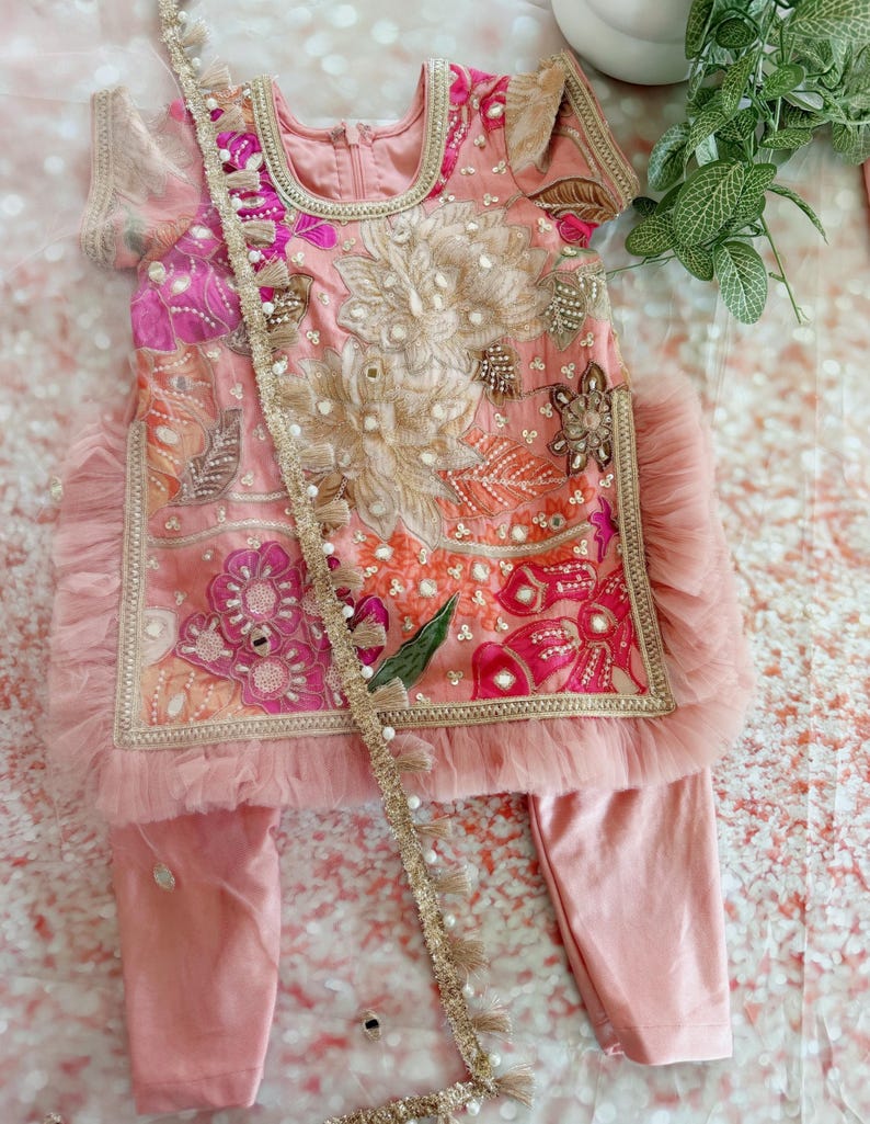 May include: A pink embroidered outfit for a child, featuring a top and pants. The top has intricate floral embroidery in shades of pink, orange, and gold, with a ruffled trim. A decorative trim with tassels and pearls accents the outfit.
